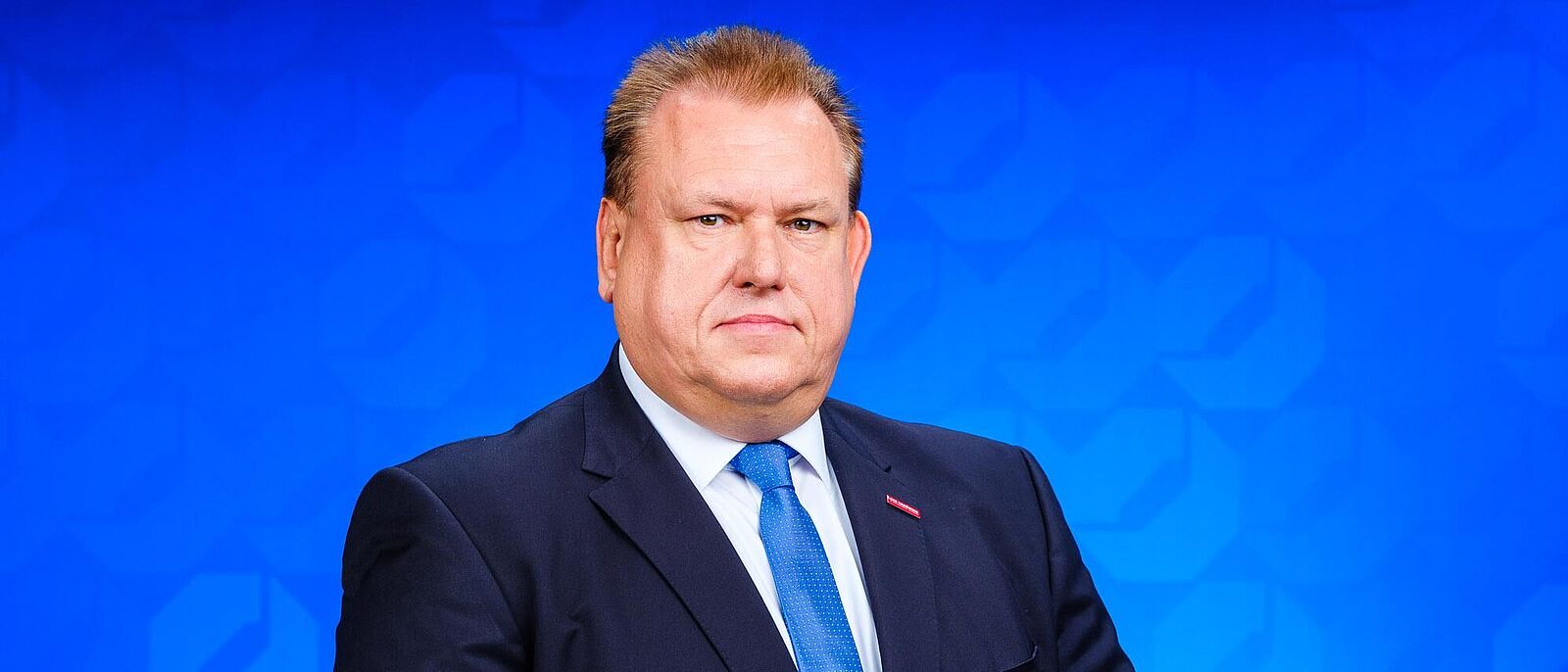 Dr. Andreas Brzezinski - Handwerkskammer Dresden Porträt von Dr. Andreas Brzezinski welcher mit aufeinandergelegten Händen vor einer blauen Logo-Wand steht.