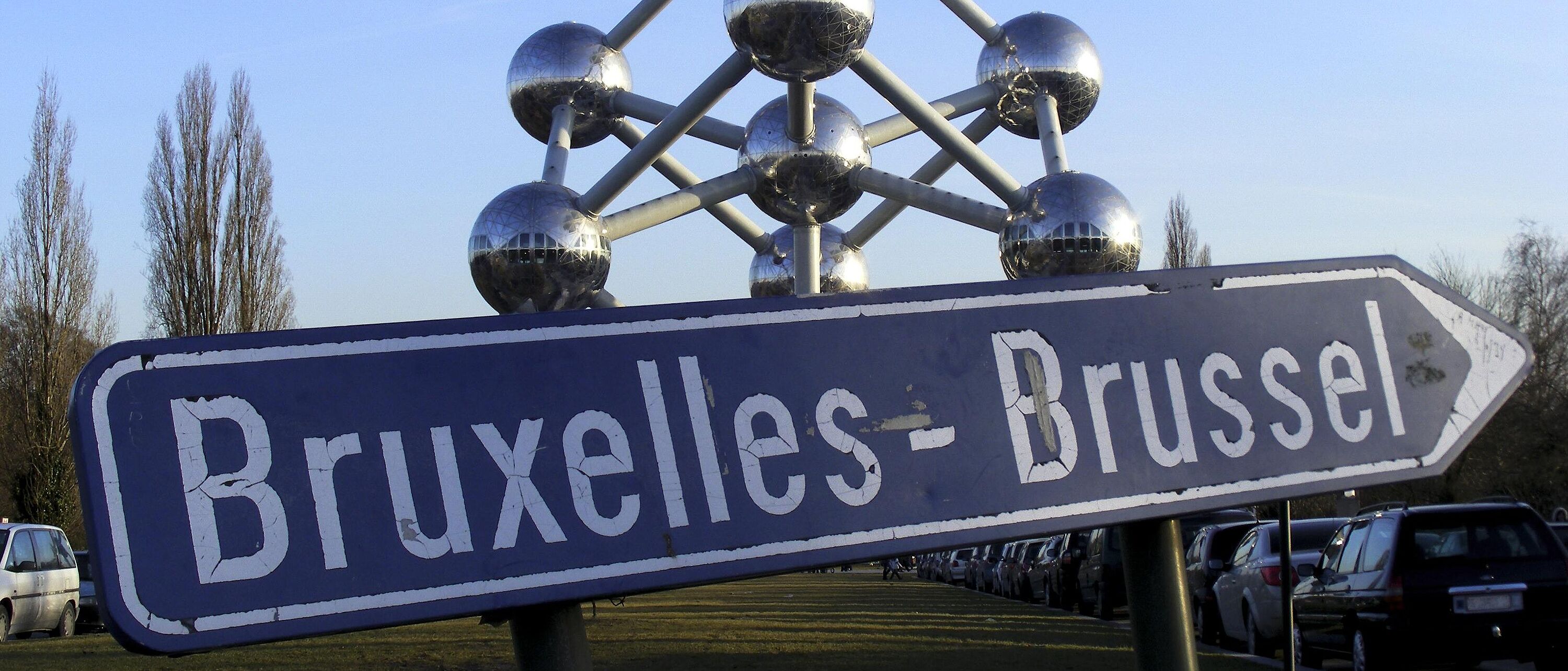 Fotografie eines blaue Straßenschildes mit der Aufschrift \"Bruxelles – Brussel\". Im Hintergrund eine Wiese und parkende Autos.