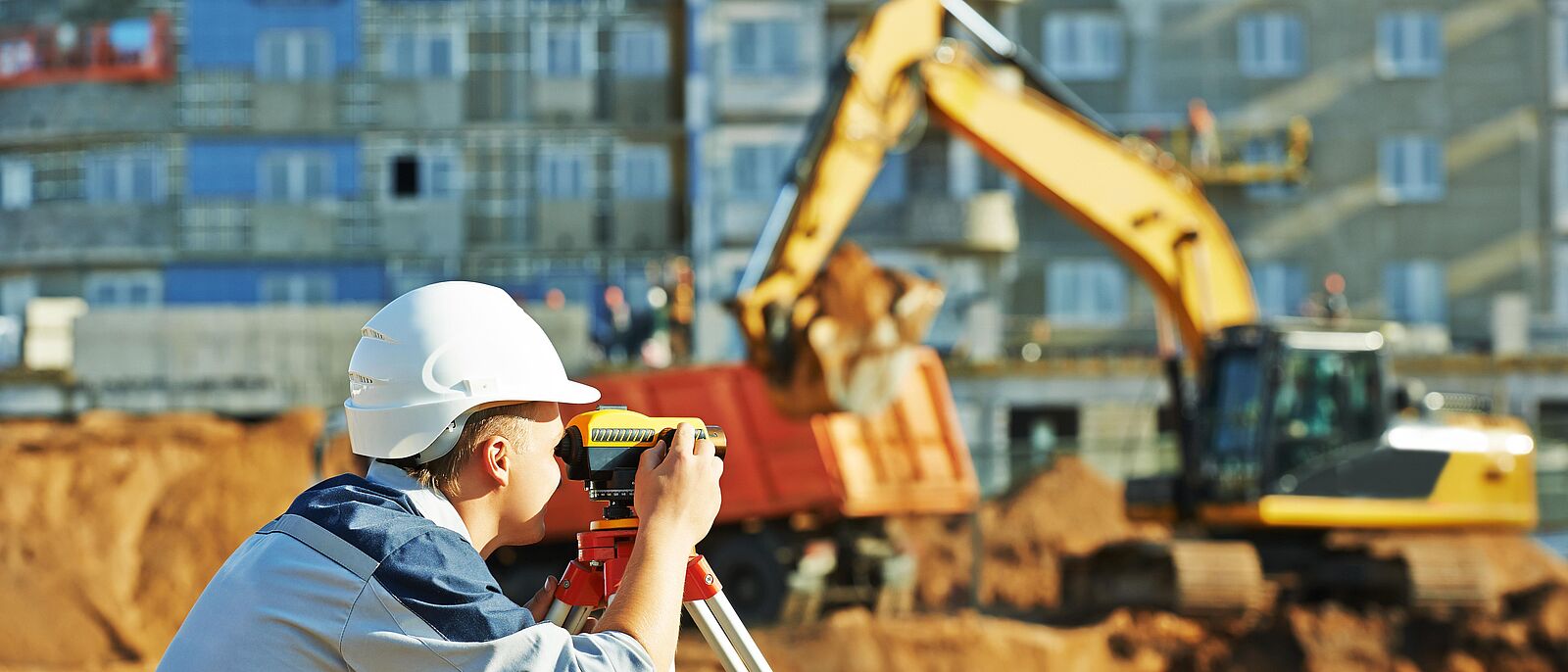 Eine Person mit einem Theodolit arbeitet auf einer Baustelle. Im Hintergrund ein Bagger und ein Rohbau von einem Hochhaus.