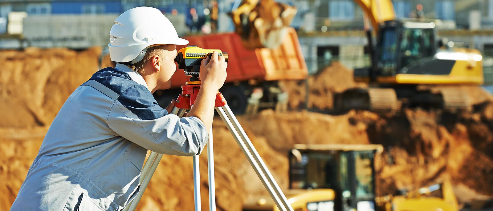 Eine Person mit einem Theodolit arbeitet auf einer Baustelle. Im Hintergrund ein Bagger und ein Rohbau von einem Hochhaus.