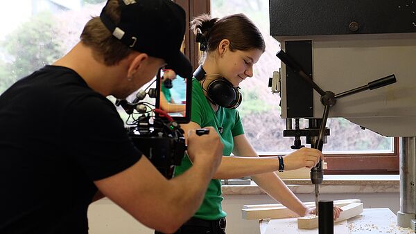 Eine Auszubildende steht an einer Bohrmaschine und wird bei ihrer konzentrierten Arbeit gefilmt und interviewt.