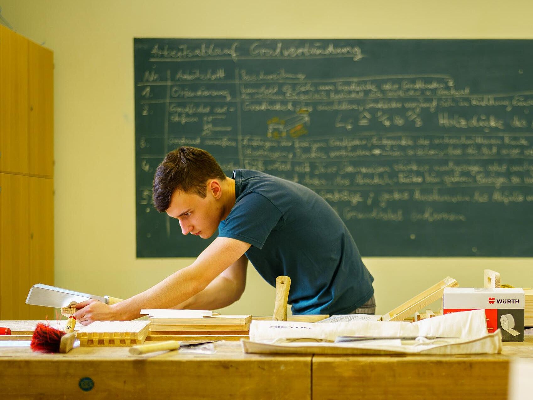 Eine Person arbeitet schleifend und prüfend an einer Werkbank in einem Tischlerraum, im Hintergrund eine voll geschriebene Unterrichtstafel.
