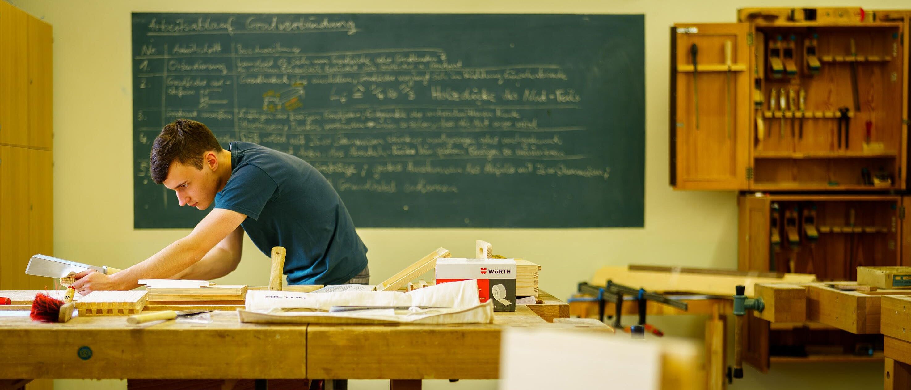 Eine Person arbeitet schleifend und prüfend an einer Werkbank in einem Tischlerraum, im Hintergrund eine voll geschriebene Unterrichtstafel.